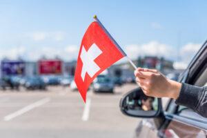 Junge hält Schweizer Flagge aus dem offenen Autofenster auf dem Parkplatz des Einkaufszentrums.