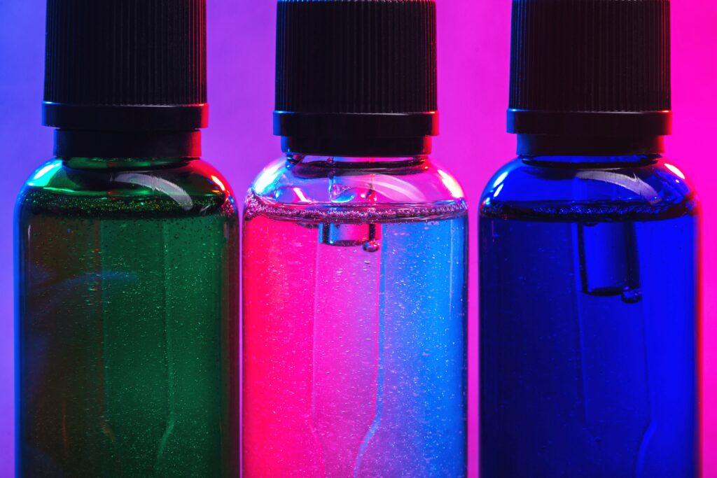 Nahaufnahme von drei E-Liquid-Flaschen in den Farben Grün, Klar und Blau, beleuchtet von einem violett-roten Hintergrund.