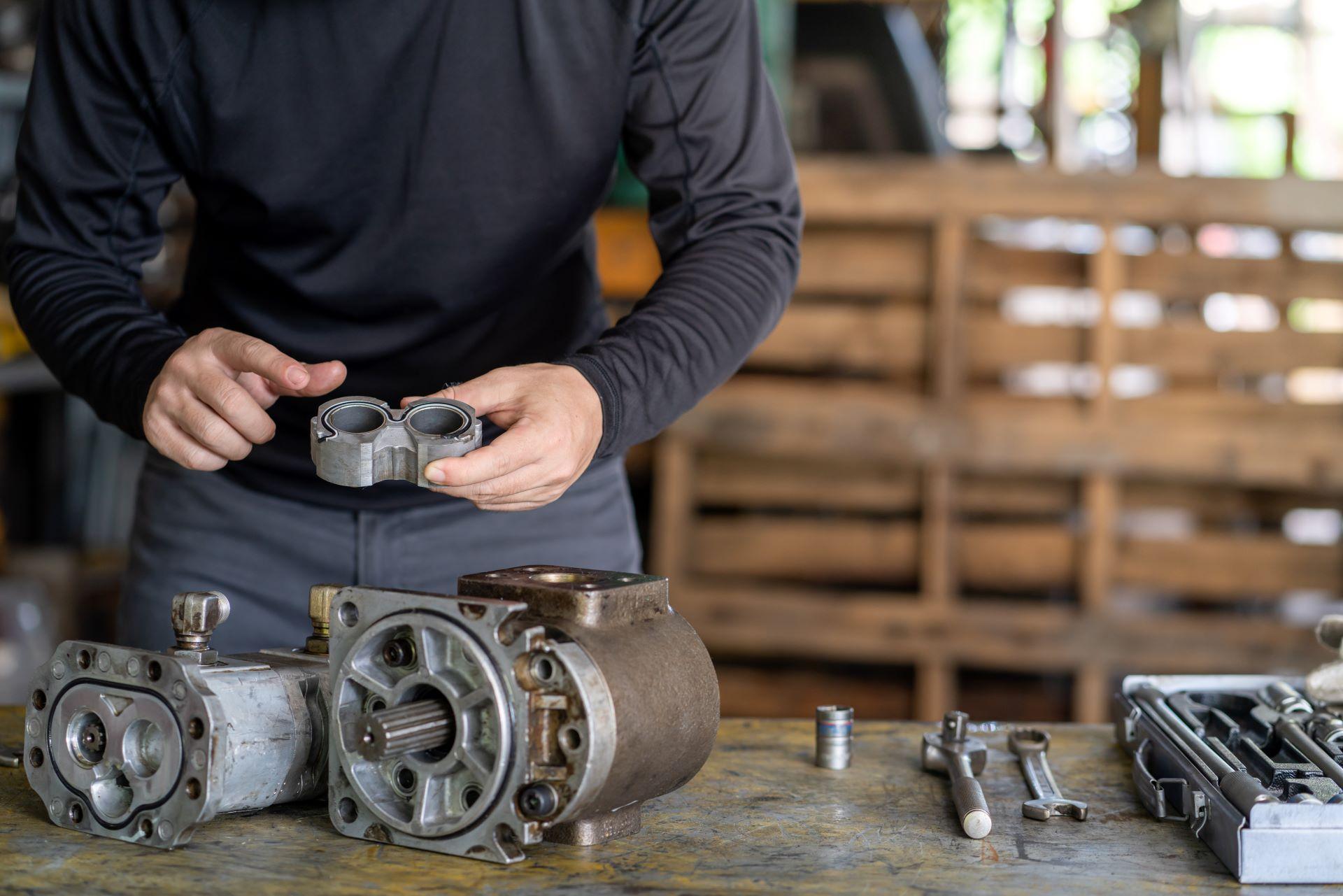 Eine Person im schwarzen Langarmshirt hält ein Bauteil einer Zahnradpumpe, während auf dem Tisch vor ihr mehrere mechanische Komponenten und Werkzeuge für die Montage oder Wartung der Pumpe liegen. Im Hintergrund ist eine Werkstatt mit Holzpaletten zu sehen.