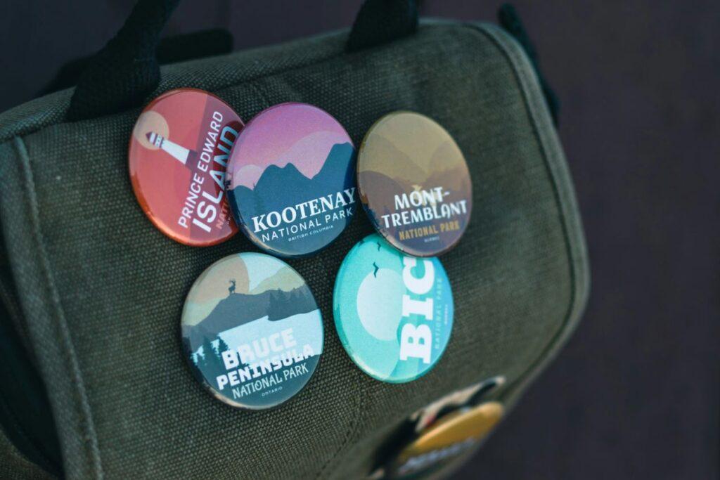 Fünf selbstgemachte Buttons mit Nationalpark-Motiven, darunter "Prince Edward Island" und "Kootenay National Park", befestigt an einer grünen Tasche.