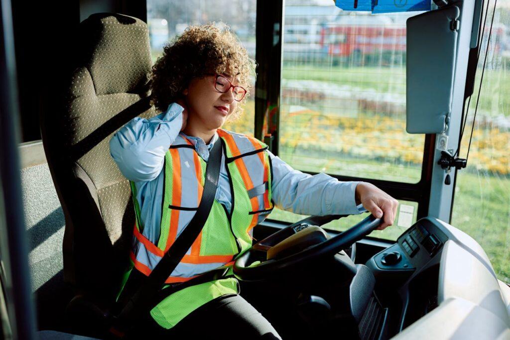 Busfahrerin mit Warnweste hält sich den Nacken vor Schmerzen. Szene aus dem Fahrerhaus, die die Bedeutung von ergonomischen Tipps und CZV-Kursen für Berufskraftfahrer*innen verdeutlicht