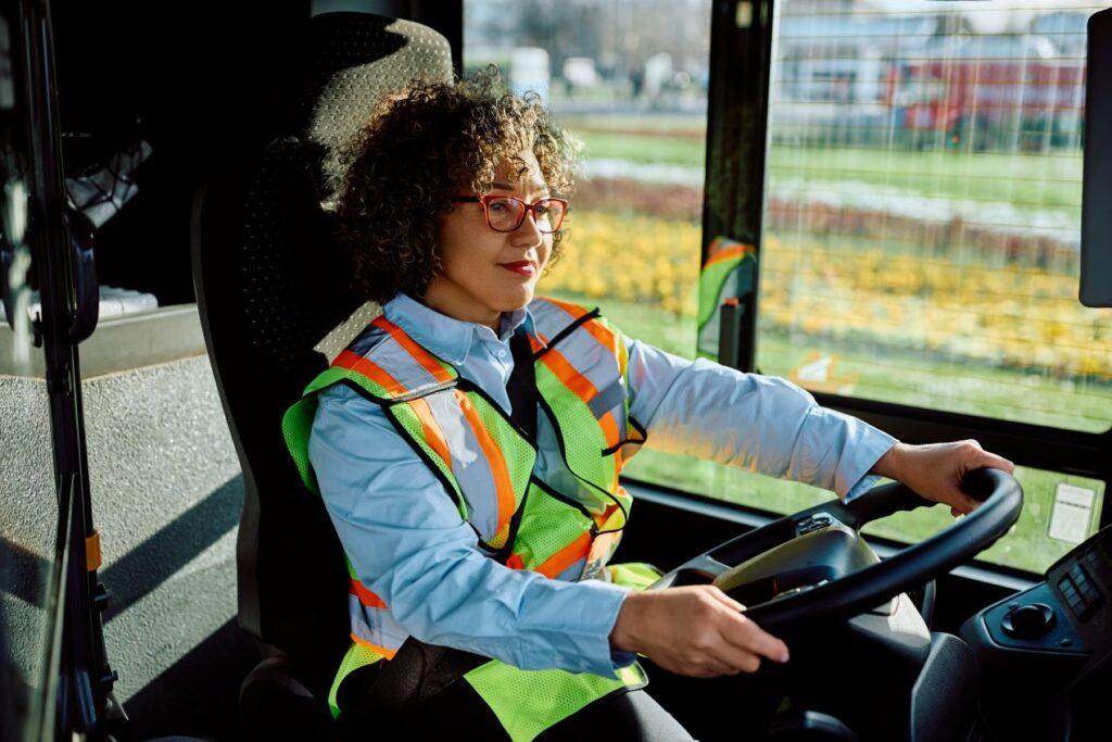 Busfahrerin mit Warnweste sitzt entspannt und aufrecht am Steuer. Beispiel für eine ergonomische Sitzhaltung, die in CZV-Kursen für Berufskraftfahrer*innen vermittelt wird