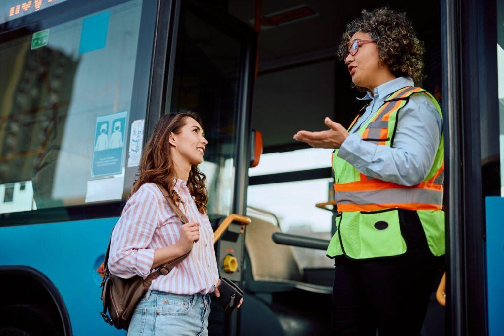 Busfahrerin in Warnweste spricht entspannt mit einer Passagierin vor ihrem Bus. Symbolbild für den Abschluss eines Arbeitstags und die Bedeutung von CZV Kursen, um Beruf und Freizeit besser zu trennen