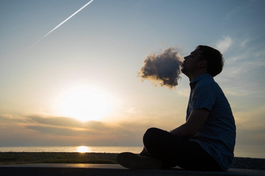 Ein Mann sitzt entspannt draußen im Sonnenuntergang und atmet eine dichte Dampfwolke aus. Der Himmel zeigt warme Farben, während die Sonne tief am Horizont steht. Die ruhige Szenerie am Wasser vermittelt ein Gefühl von Frieden und Auszeit vom Alltag.