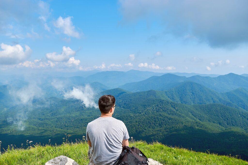 Ein Mann sitzt mit dem Rücken zur Kamera auf einem Berggipfel und schaut auf eine beeindruckende Landschaft mit grünen, bewaldeten Hügeln und einem weiten blauen Himmel. Neben ihm liegt ein Rucksack. Eine Dampfwolke steigt neben ihm auf, während er die Aussicht in Ruhe genießt. Die Szene strahlt Freiheit, Ruhe und Verbundenheit mit der Natur aus.