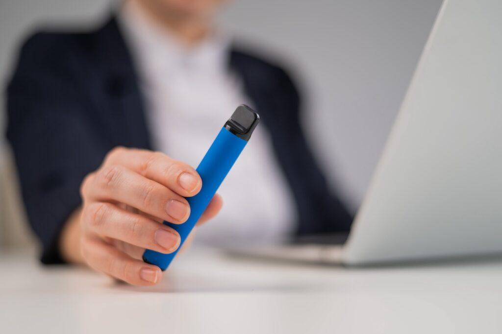 Eine Person in Business-Kleidung hält eine blaue Elf Bar Pod in der Hand. Der Fokus liegt auf der Vape, während im Hintergrund ein Laptop sichtbar ist. Das Bild symbolisiert kleine Genussmomente während der Arbeit oder im stressigen Alltag.