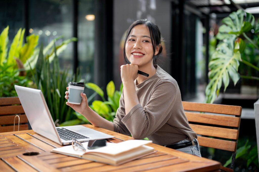 Eine asiatische Frau, die aus einem ruhigen Café heraus arbeitet, sitzt an einem Tisch im Freien mit ihrem Laptop.