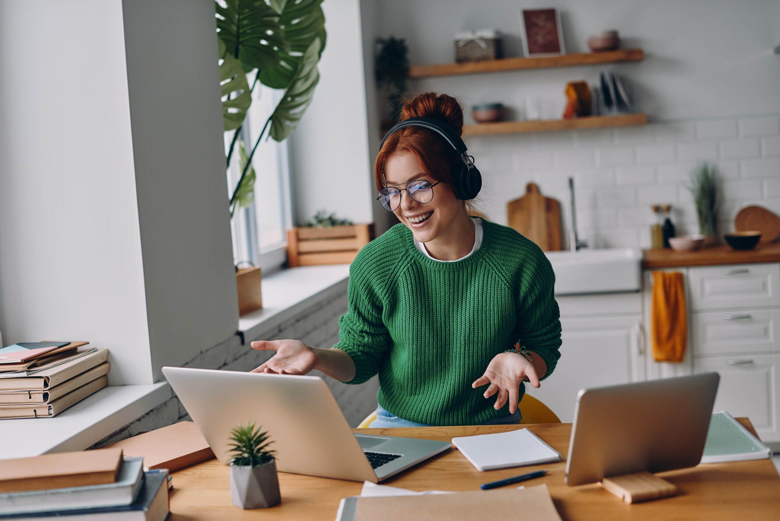 Fröhliche junge Frau mit Kopfhörern, die während der Arbeit von zu Hause aus an einer Webkonferenz teilnimmt.